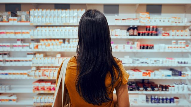 Rear view of young woman with bag standing against shelf in pharmacy searching for medicine