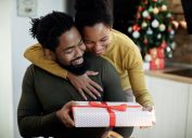 Happy black woman embracing her husband and giving his a gift on Christmas day at home.