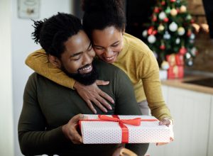 Happy black woman embracing her husband and giving his a gift on Christmas day at home.