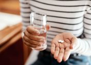 Woman in striped shirt holding glass of water in one hand and tablets in the other.