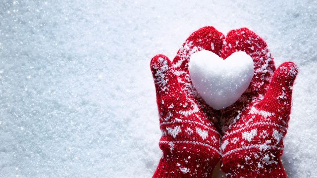 Hands wearing mittens and holding heart made out of snow.