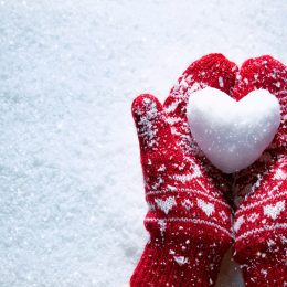 Hands wearing mittens and holding heart made out of snow.
