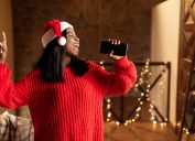 Christmas holiday music. Happy black lady in Santa hat singing, using smartphone as mic at home, mockup for mobile app. Cheerful young woman enjoying karaoke song, having fun on Xmas
