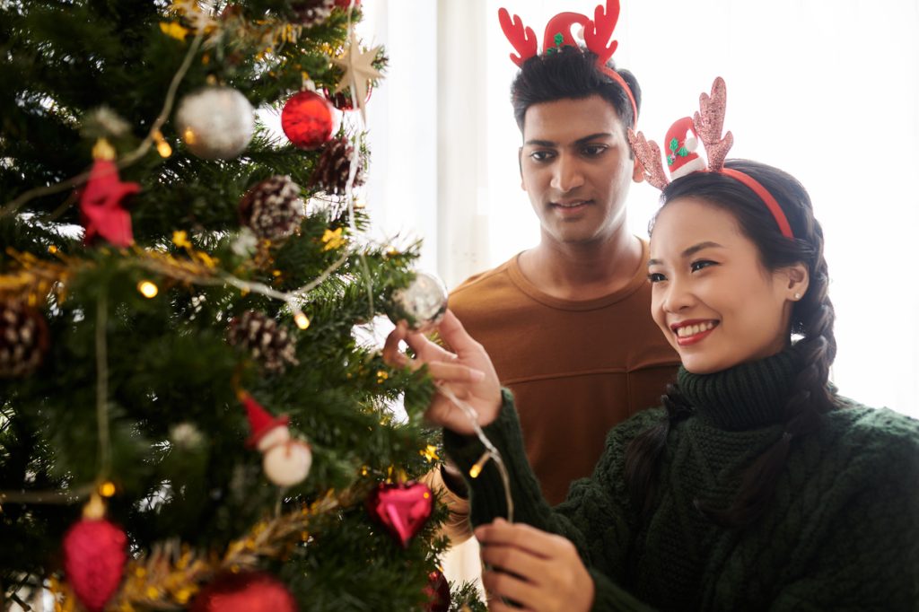 Happy devirse young couple decorating Christmas tree at home with colorful baubles