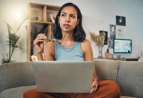 Beautiful mixed race woman thinking while using laptop for blogging in living room at home. Hispanic entrepreneur sitting cross legged alone on lounge sofa 