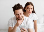 A suspicious-looking woman looking over her male partner's shoulder at his text messages.