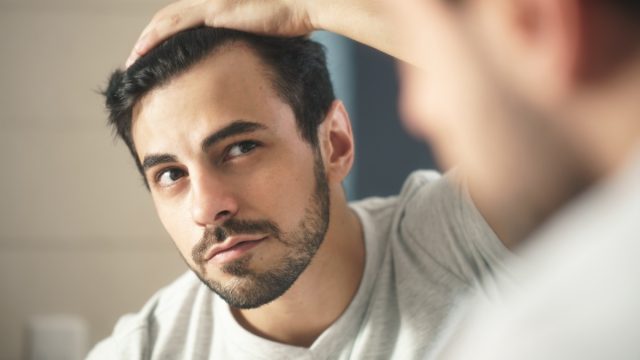 person with beard grooming in bathroom at home. White metrosexual man worried for hair loss and looking at mirror his receding hairline.