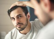 người đàn ông với bộ râu chải chuốt trong phòng tắm ở nhà.  Người đàn ông chuyển giới da trắng lo lắng về chứng rụng tóc và nhìn vào gương để thấy chân tóc ngày càng thưa dần của mình.