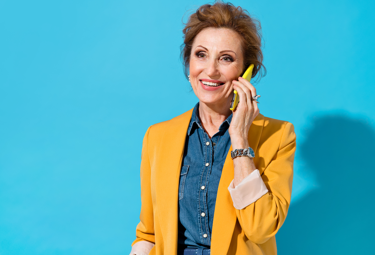 An older woman with auburn hair wearing a denim shirt and mustard-yellow blazer talks on the phone against a teal background.