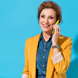 An older woman with auburn hair wearing a denim shirt and mustard-yellow blazer talks on the phone against a teal background.