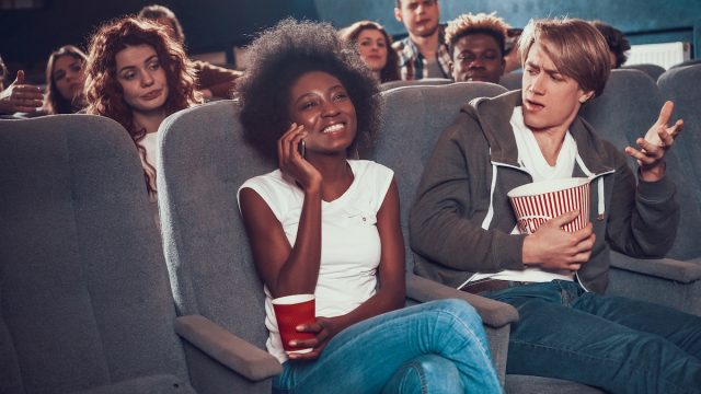 A young woman smiling and talking on the phone in the movie theater, while everyone around her looks annoyed.