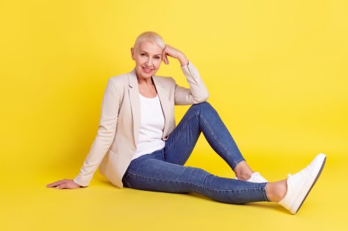 An older woman sitting on a yellow background, wearing jeans and a beige blazer.