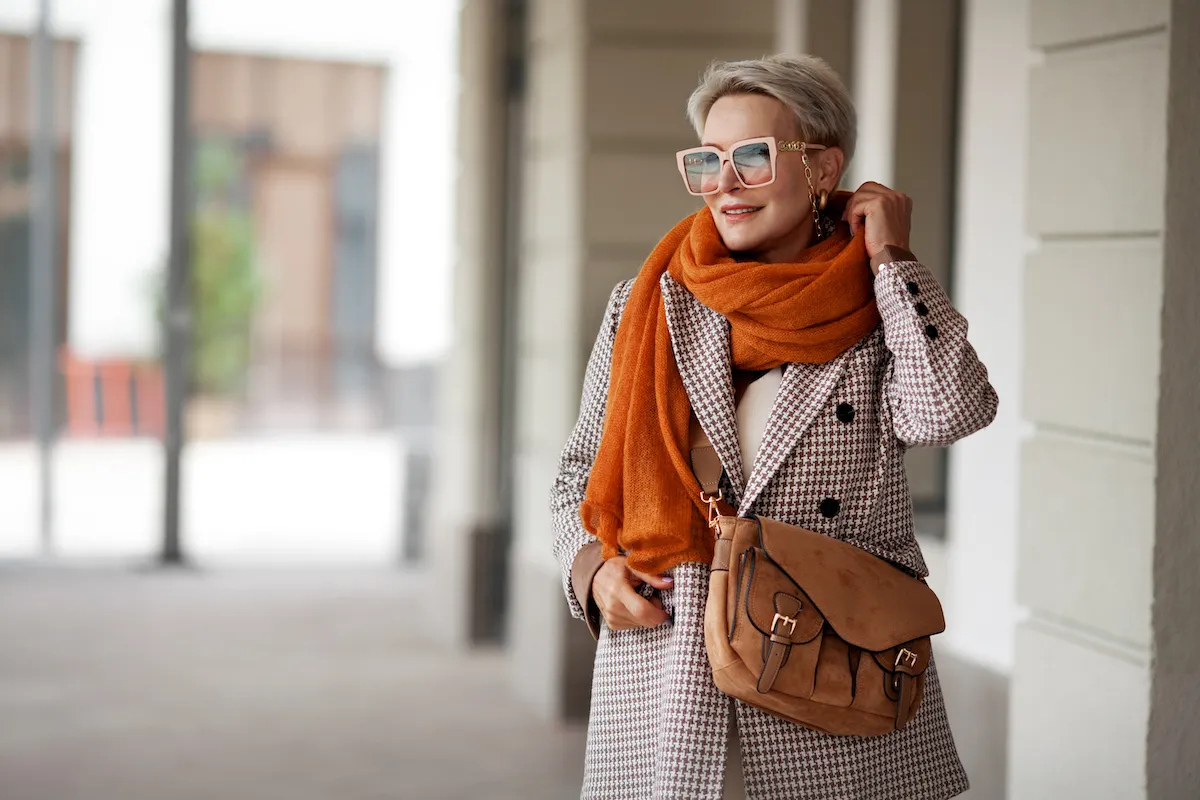 Smiling Woman Outdoor Portrait. Short blonde hair fashion model wears stylish clothes, double-breasted jacket, leather handbag, ochre knitted scarf and glasses. Fashion trend of autumn or spring