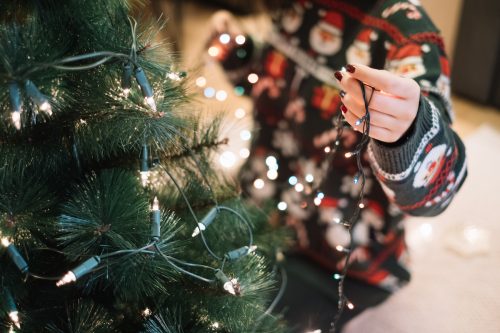 Close up of a woman in a Santa sweater putting lights on her Christmas tree.