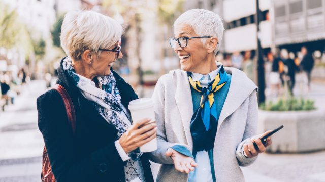 Two senior women, both with short white hair and wearing jackets and scarves, walk down the street laughing.