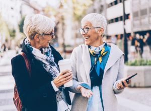Two senior women, both with short white hair and wearing jackets and scarves, walk down the street laughing.