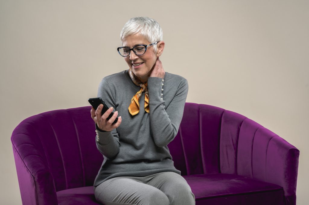 stylish Older woman with short hair on purple couch looks at phone