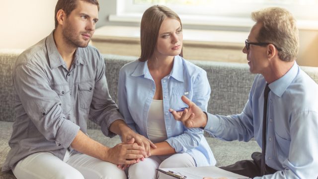A young couple holding hands shares the couch with their therapist who's talking to them.