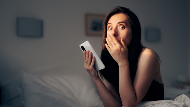 A shocked-looking young woman in bed holding her phone.