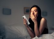 A shocked-looking young woman in bed holding her phone.