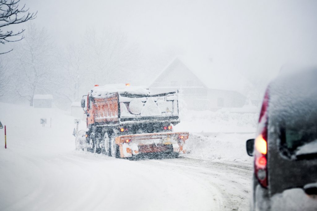A car driving behind a snow plow on a snowy road during a blizzard