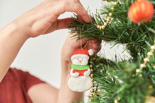 Unrecognizable woman decorating a Christmas tree at home, close up of the hands hanging a snowman ornament on a branch.