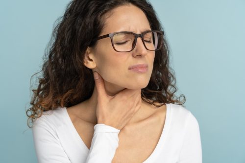 Woman having sore throat, tonsillitis, feeling sick, suffering from painful swallowing, angina, strong pain in throat, loss of voice, holding hand on her neck, isolated on studio blue background.