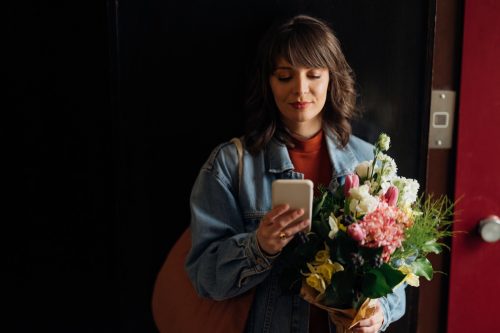 Smiling woman typing text message on her smartphone while holding bouquet of flowers and standing at the elevator in the building.
