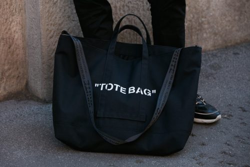 Close up of a black tote bag at a man's feet with the words "tote bag" written in white letters.