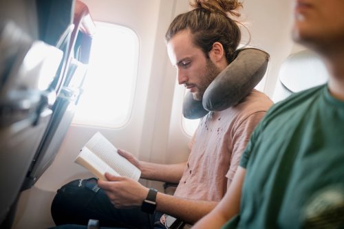 One young man is reading a book while travelling on an airplane. He is using a neck pillow.