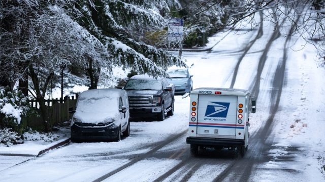 United States Postal Service (USPS) delivery truck driving down a snowy residential street during a winter storm, with space for text