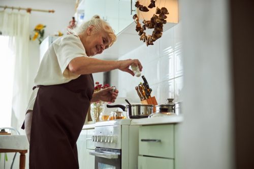 Cheerful old lady adding salt to soup and smiling while standing by the stove with cooking pots