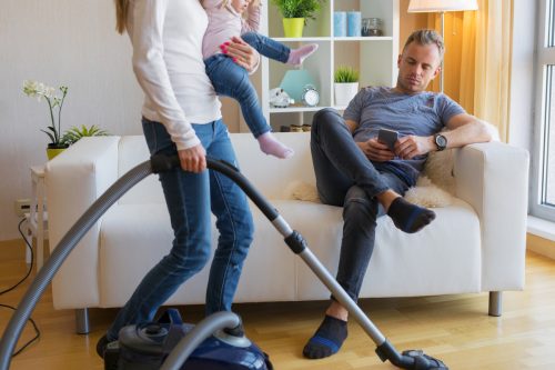 A woman holding a small child is vacuuming while her husband sits on the couch on his phone.