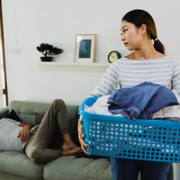 An annoyed-looking wife carries the laundry basket while her husband sleeps on the couch.
