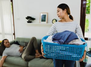An annoyed-looking wife carries the laundry basket while her husband sleeps on the couch.