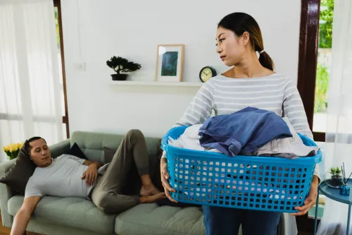 An annoyed-looking wife carries the laundry basket while her husband sleeps on the couch.