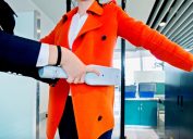 Close up of a woman in a bright orange coat being scanned with the metal detector at airport security.