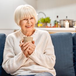 A smiling senior woman wearing a cozy sweater and holding a cup of tea on her couch.