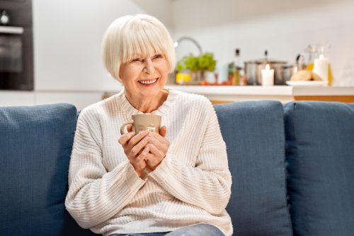 A smiling senior woman wearing a cozy sweater and holding a cup of tea on her couch.