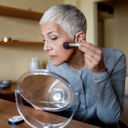 A woman with short gray hair wearing a gray cardigan puts makeup on in a small mirror.