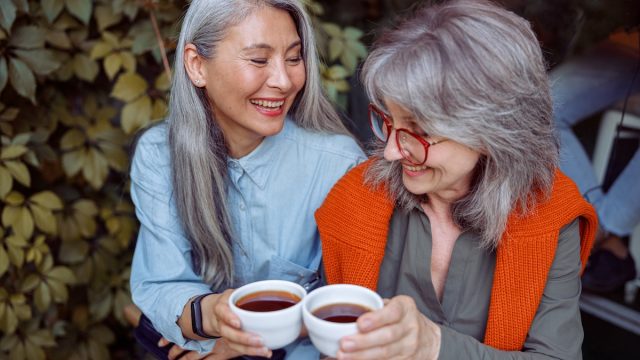 Two women friends with long gray hair having coffee and smiling.
