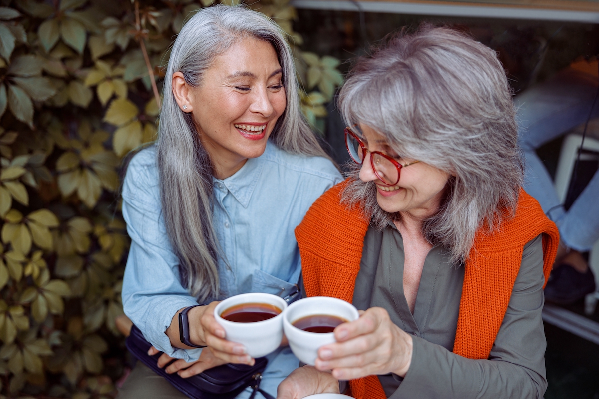 Hai người phụ nữ thân thiện với mái tóc hoa râm dài đang uống cà phê và mỉm cười.