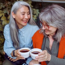 Two women friends with long gray hair having coffee and smiling.