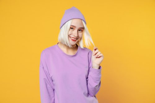 Smiling young blonde woman wearing a basic purple shirt and matching hat looking at the camera playing with her hair on an isolated yellow background. 