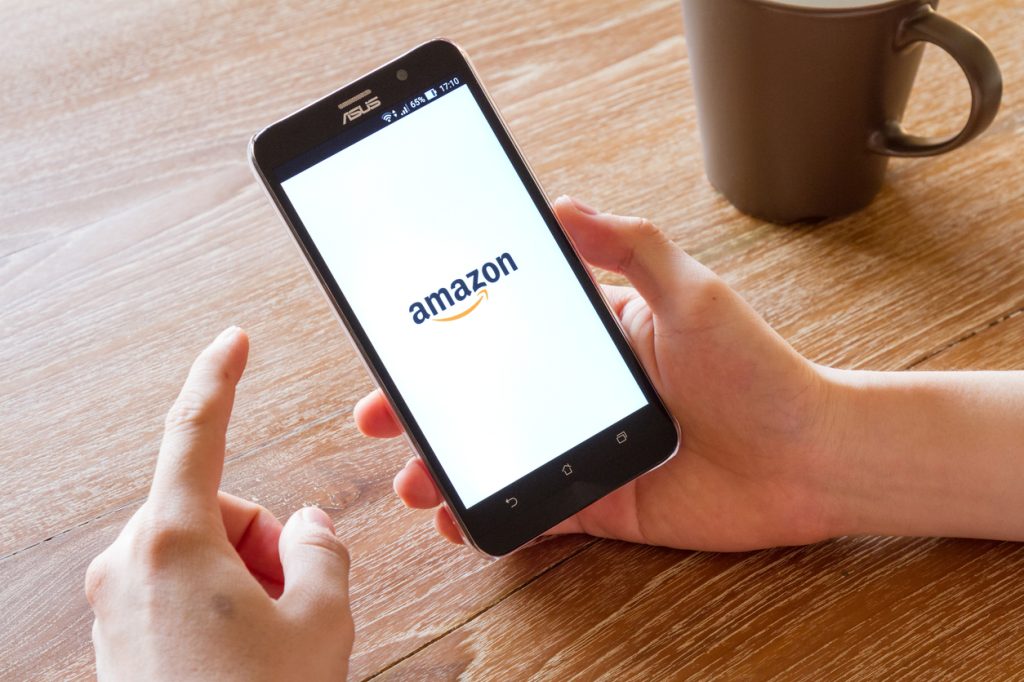 A close up of the Amazon logo on a smartphone in someone's hand next to a coffee mug