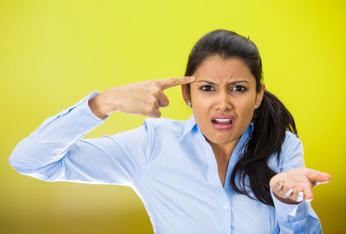 Closeup portrait of angry mad young woman gesturing with her finger against temple asking are you crazy? She's wearing a light-blue shirt against a bright yellow background.