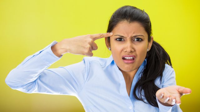 Closeup portrait of angry mad young woman gesturing with her finger against temple asking are you crazy? She's wearing a light-blue shirt against a bright yellow background.