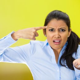 Closeup portrait of angry mad young woman gesturing with her finger against temple asking are you crazy? She's wearing a light-blue shirt against a bright yellow background.