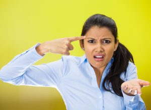 Closeup portrait of angry mad young woman gesturing with her finger against temple asking are you crazy? She's wearing a light-blue shirt against a bright yellow background.