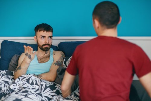 Young man in bed looking at his boyfriend and explaining, using hand gesture, LGBT break up